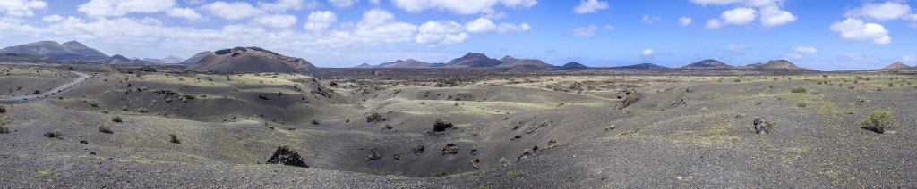 Ein Wanderweg in der Nähe des Timanfaya Nationalparks führt einmal rund um die Caldera de los Cuervos. Eine Explosion hat den Krater an einer Seite geöffnet (siehe den heraus geschleuderten Felsblock im Lavafeld rechts neben der Caldera), so dass man durch die Explosionsöffnung ins innere des Kraters gelangen kann, Lanzarote, Kanarische Inseln, März 2017.