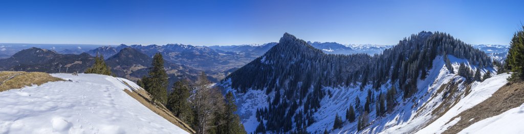 Panorama vom Gipfel der Brünnsteinschanze (1547m) gegen Osten mit Blick auf die Chiemgauer Alpen, das Gebiet um den Walchsee, Zahmen und Wilden Kaiser, zum Teil verdeckt durch den Gipfel des Brünnsteins (1634m) sowie die Rotwandlspitz (1588m). Dazwischen reicht die Fernsicht bis zum fernen Großglockner in den Hohen Tauern, Mangfallgebirge, Bayrische Alpen, Februar 2021.