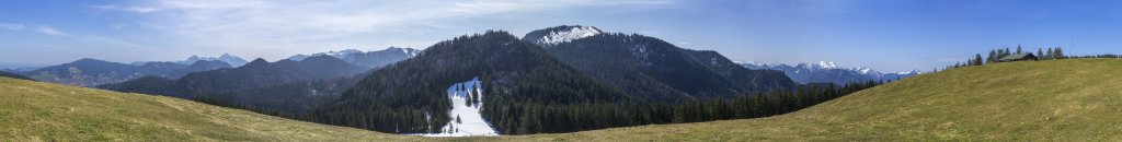 Panormablick am Kreuz der Kreuzbergalm (1222m) mit Blick auf den Schliersberg, den Wendelstein, Rotwandgebiet, das gegenüberliegende Kreuzbergköpfel (1273m) und die Baumgartenschneid (1444m) sowie die verschneiten Gipfel von Hirschberg, Ochsen-, Auer-, Spitzkampel und Fockenstein, Mangfallgebirge, Bayrische Alpen, April 2021.